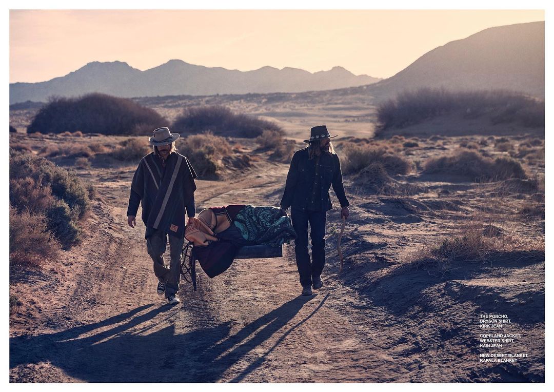 Two adult men are walking through a desert carrying a covered stretcher, under warm lighting, with distant mountains in the background.