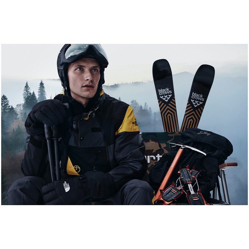 Man in ski gear posing with skis, an ice axe, and a backpack against a mountain backdrop with trees and a foggy sky. Winter sport apparel.
