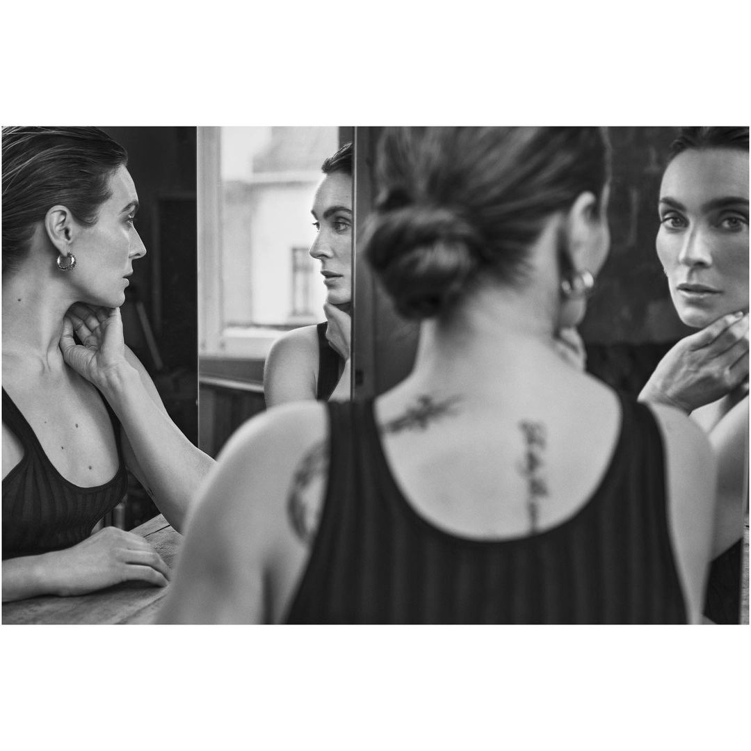 Monochrome shot of a woman with slicked-back hair and earrings, looking at herself in a triple mirror, hand to her neck indoors.