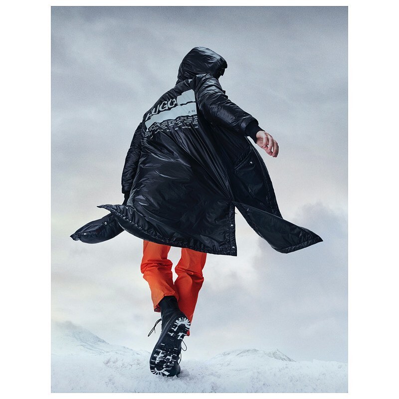 Person in a black puffer coat and orange pants walks in the snow, showcasing the back of the coat with a white logo against a cloudy sky.