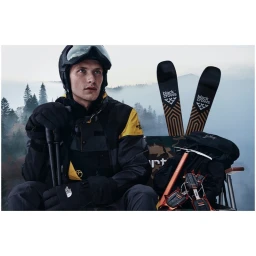 Male skier in black and yellow jacket and ski goggles holding poles with skis and gear in the background
