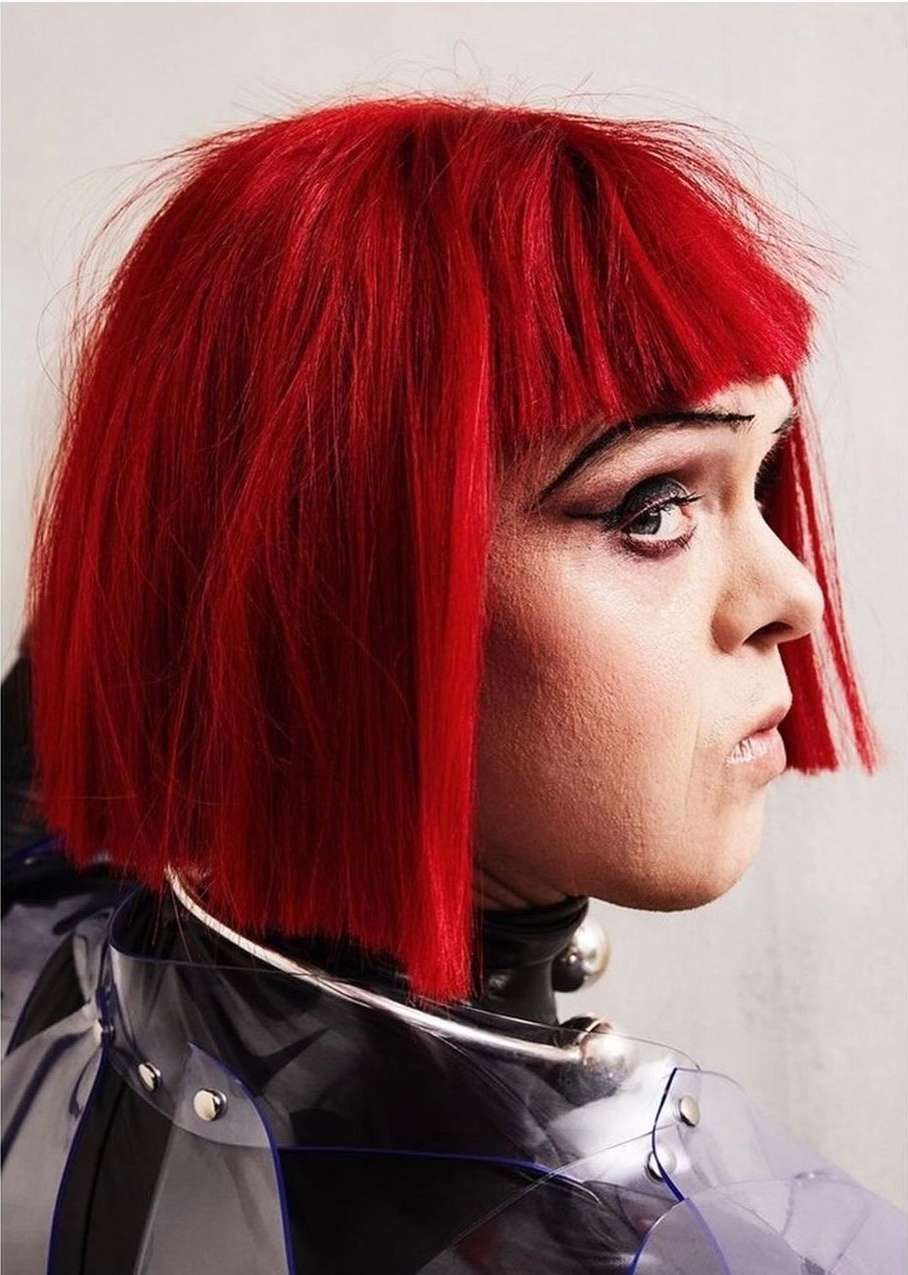 Close-up portrait of a person with vibrant red bob haircut and striking eye makeup, wearing a transparent, studded raincoat and silver neck ring.