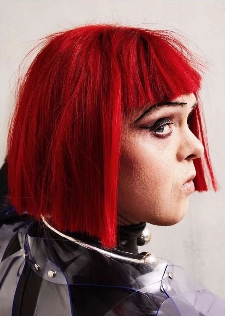 Close-up portrait of a person with vibrant red bob haircut and striking eye makeup, wearing a transparent, studded raincoat and silver neck ring.