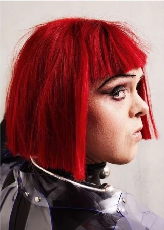 Close-up portrait of a person with vibrant red bob haircut and striking eye makeup, wearing a transparent, studded raincoat and silver neck ring.