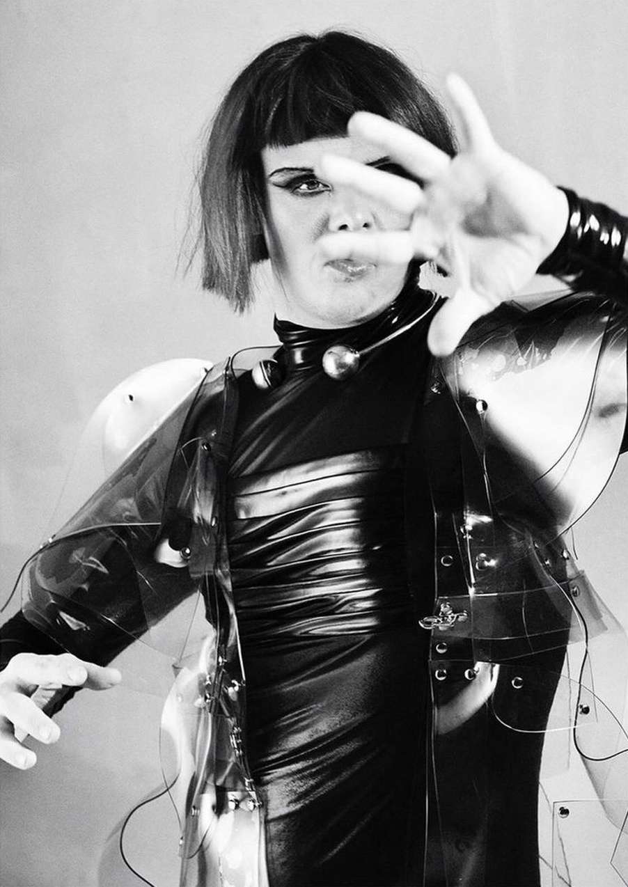 Monochrome portrait of a non-binary person with makeup and a clear plastic garment holding up a hand with fingers spread in front of face.
