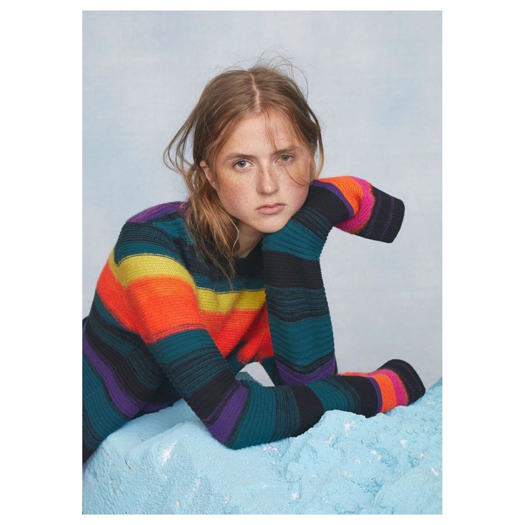 Young woman with freckles gazes at the camera, wearing a striped knit sweater and resting her arms on a blue, textured surface.