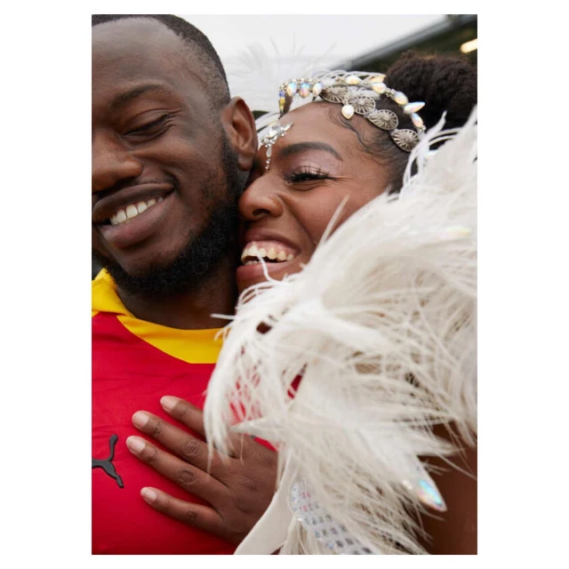 A joyful couple, an adult man and woman, embrace and smile broadly, the woman in a feathered costume and the man in a Ghana shirt.