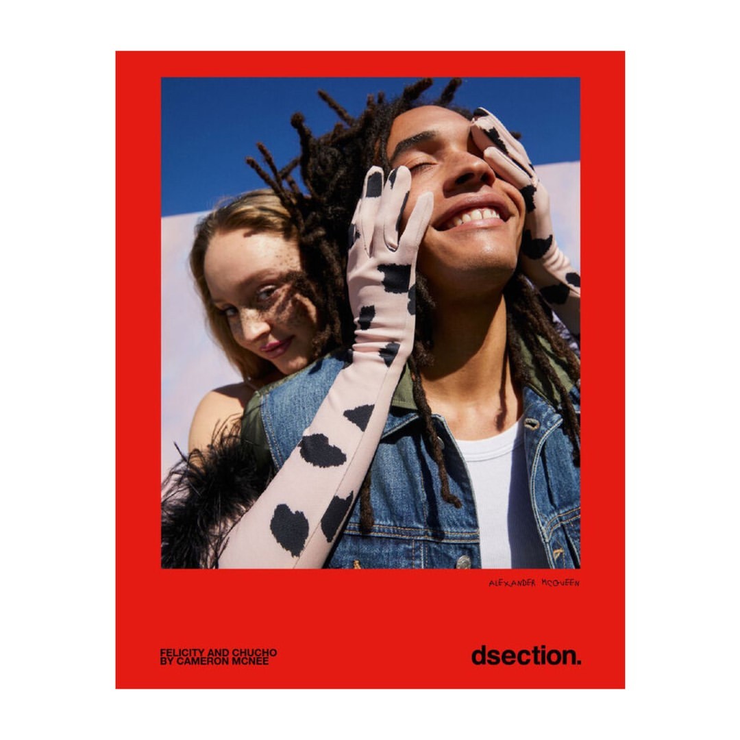 Fashion editorial featuring a smiling man with dreadlocks and gloves with a woman with blonde hair, set against a red and blue background.