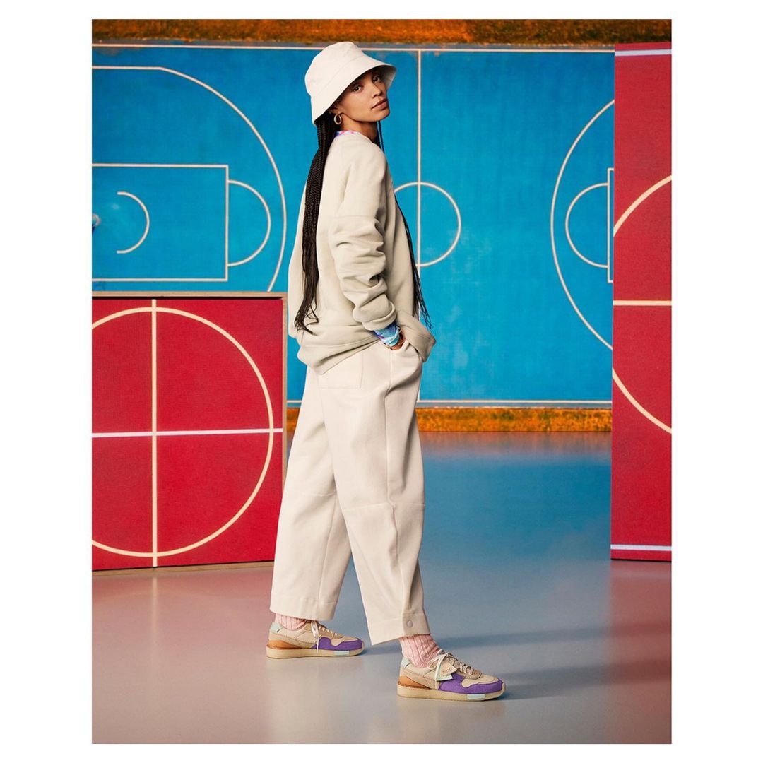 Woman in bucket hat and casual wear poses in sneakers in front of a colorful basketball court backdrop inside of a photo studio.