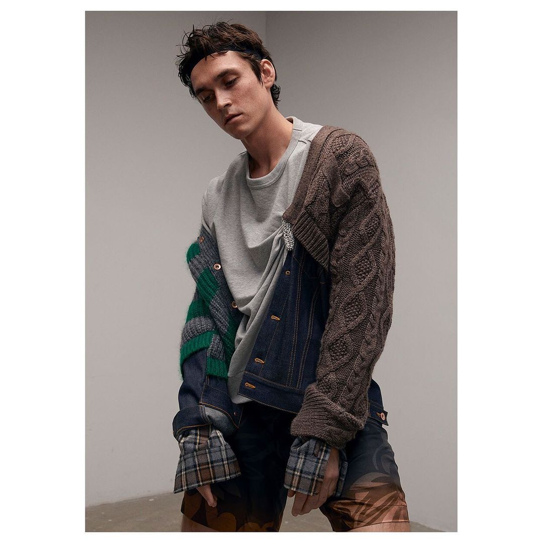 A young adult male poses in a studio wearing a gray t-shirt and a deconstructed, multi-textured jacket against a neutral background.