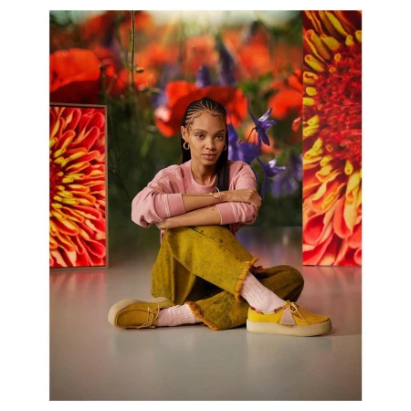 A young adult female model sits in a studio with colorful flower backdrops, wearing a pink top, yellow pants and matching yellow platform shoes.