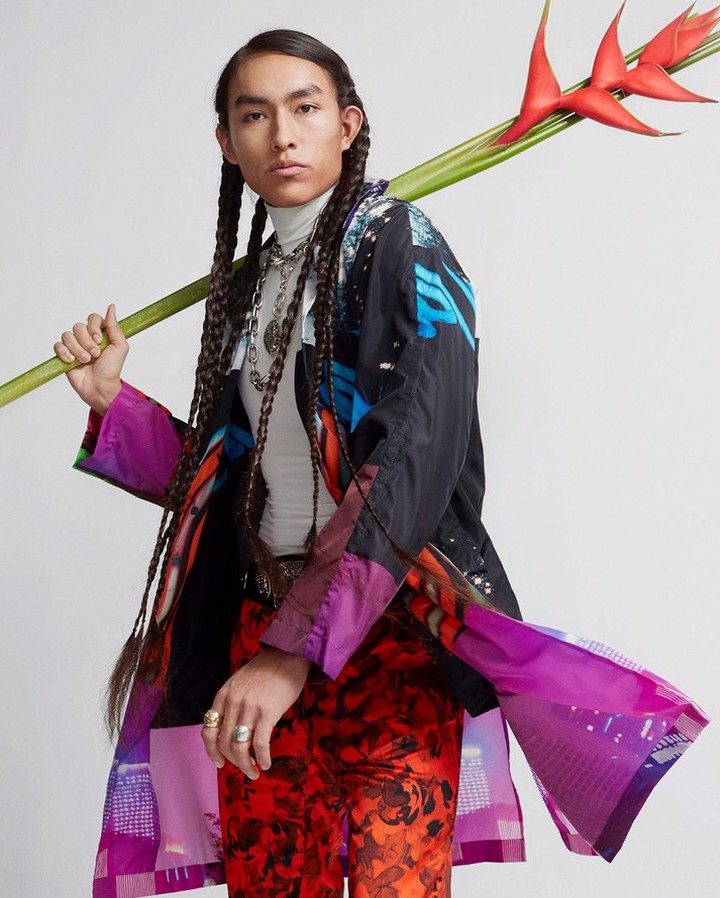 Portrait of a young adult male with long braided hair wearing a patterned coat, red pants, and holding a tropical flower. Studio shot.