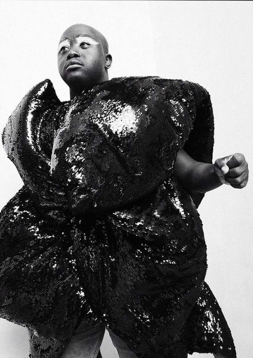 Striking black and white studio portrait of bald African American person in a large, sculptural, shiny black sequin garment.