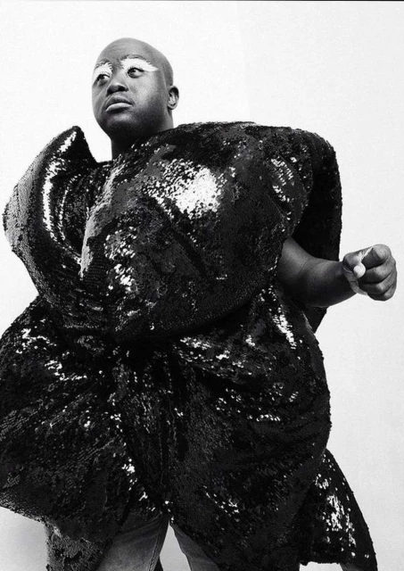 Striking black and white studio portrait of bald African American person in a large, sculptural, shiny black sequin garment.