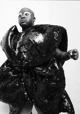 Striking black and white studio portrait of bald African American person in a large, sculptural, shiny black sequin garment.