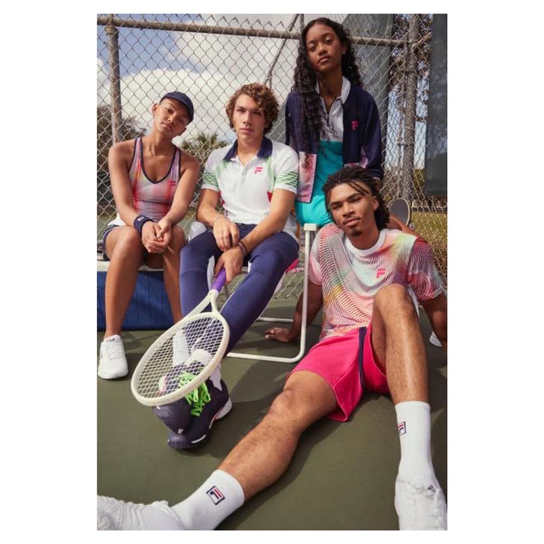 Four young adults in athleisure wear pose near a tennis court with rackets. The diverse group is clad in colorful sportswear by FILA.