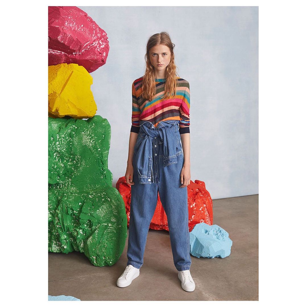 An adult female poses in a multicolored striped top and denim pants, accessorized with white sneakers, in a studio setting with colorful rock props.