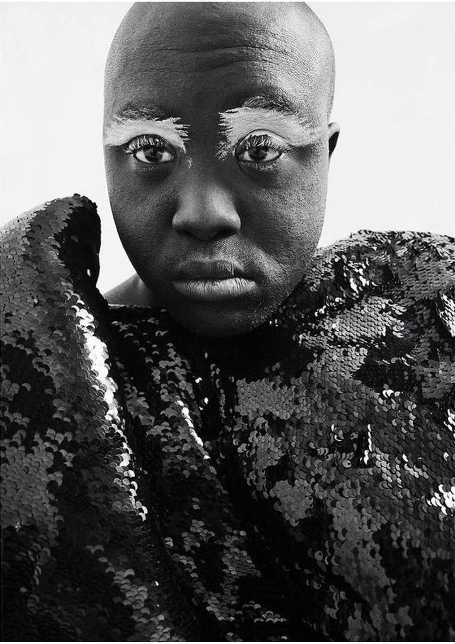 Close-up portrait of bald adult with white eyebrow makeup and a shiny sequin garment. The image is high-contrast black and white.