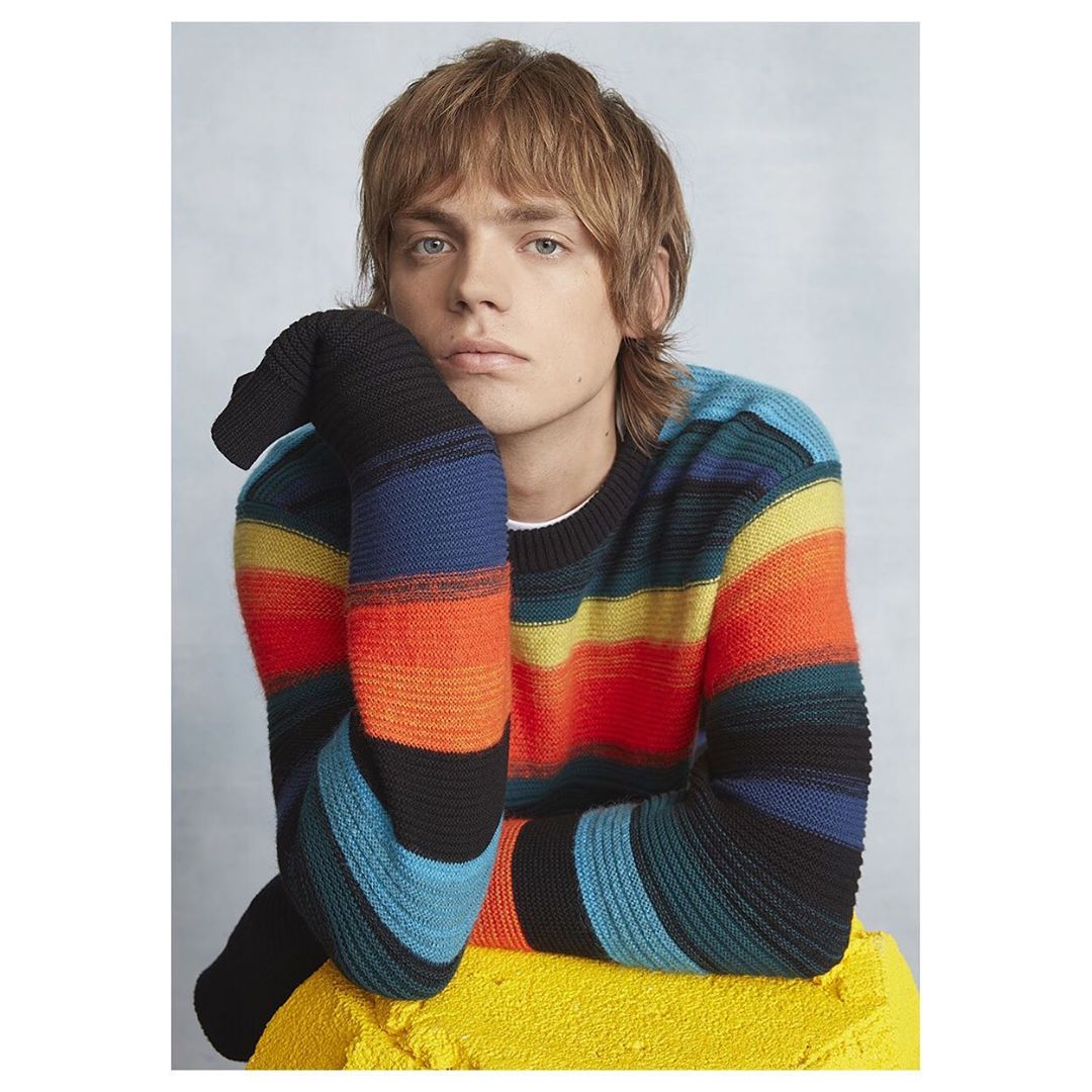 Portrait of a young adult male wearing a colorful striped sweater, resting his head on his hand, posing against a neutral background.