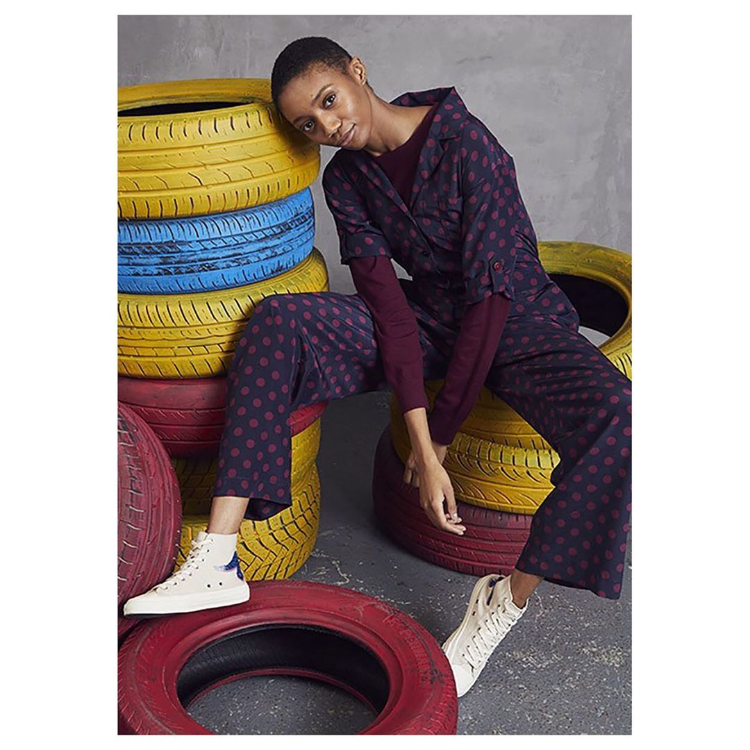 A young woman with short hair poses while sitting on a stack of colorful tires, wearing a polka dot pantsuit and white sneakers.