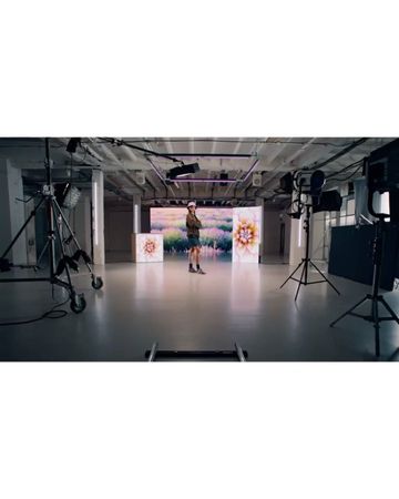 A person stands in a photo studio surrounded by lighting equipment and screens displaying floral images in a bright, spacious setting.