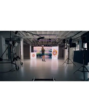 A person stands in a photo studio surrounded by lighting equipment and screens displaying floral images in a bright, spacious setting.