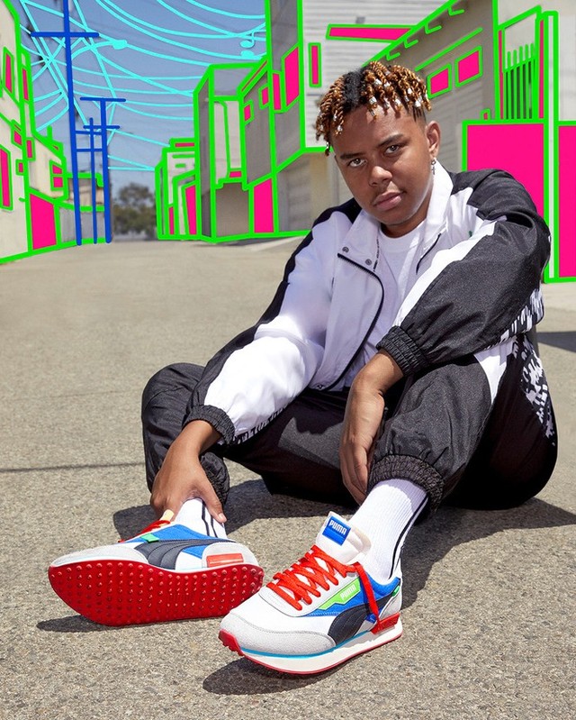 Person sitting on a road, wearing Puma jacket and pants, with Puma sneakers in foreground, against an illustrated urban background.
