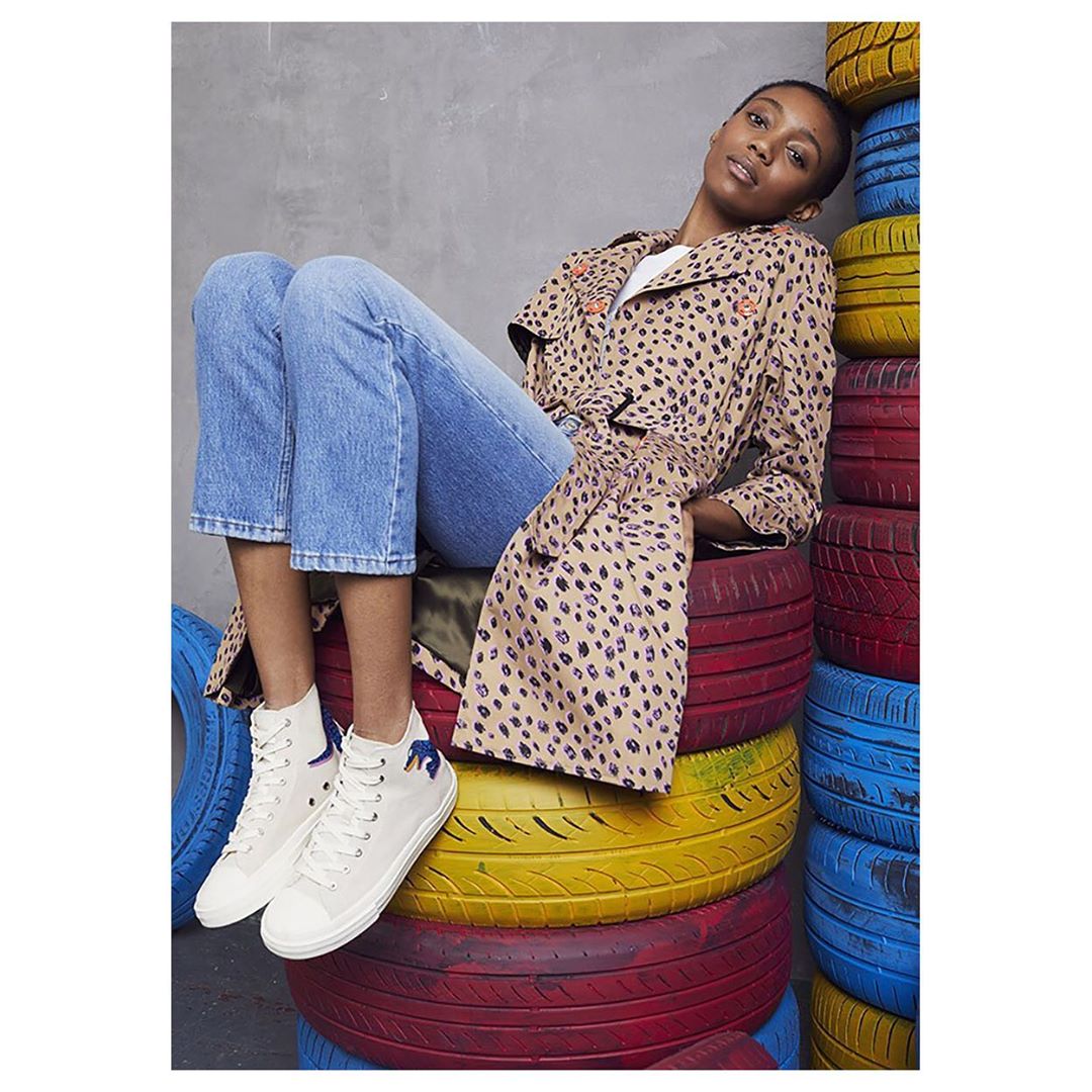 Stylish woman in a leopard-print trench coat and jeans sitting atop a stack of brightly painted tires in a studio setting.