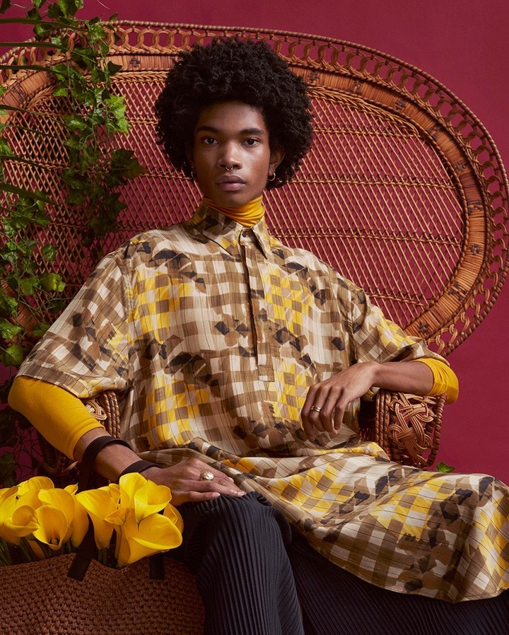 Portrait of a stylish person with afro, wearing patterned tunic, seated in a wicker chair with a maroon background and floral accent.