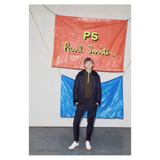 Male model wearing dark jacket and pants, standing in front of a backdrop with the 'Paul Smith' logo in an indoor setting.