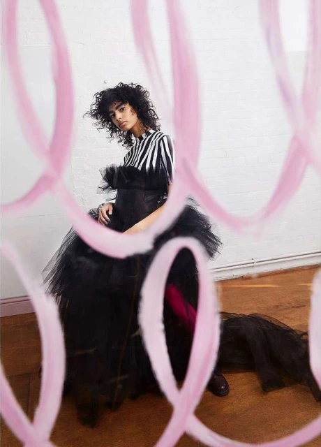 A woman wearing a black and white striped top and a black tulle dress is seen through pink abstract shapes in a photo studio.