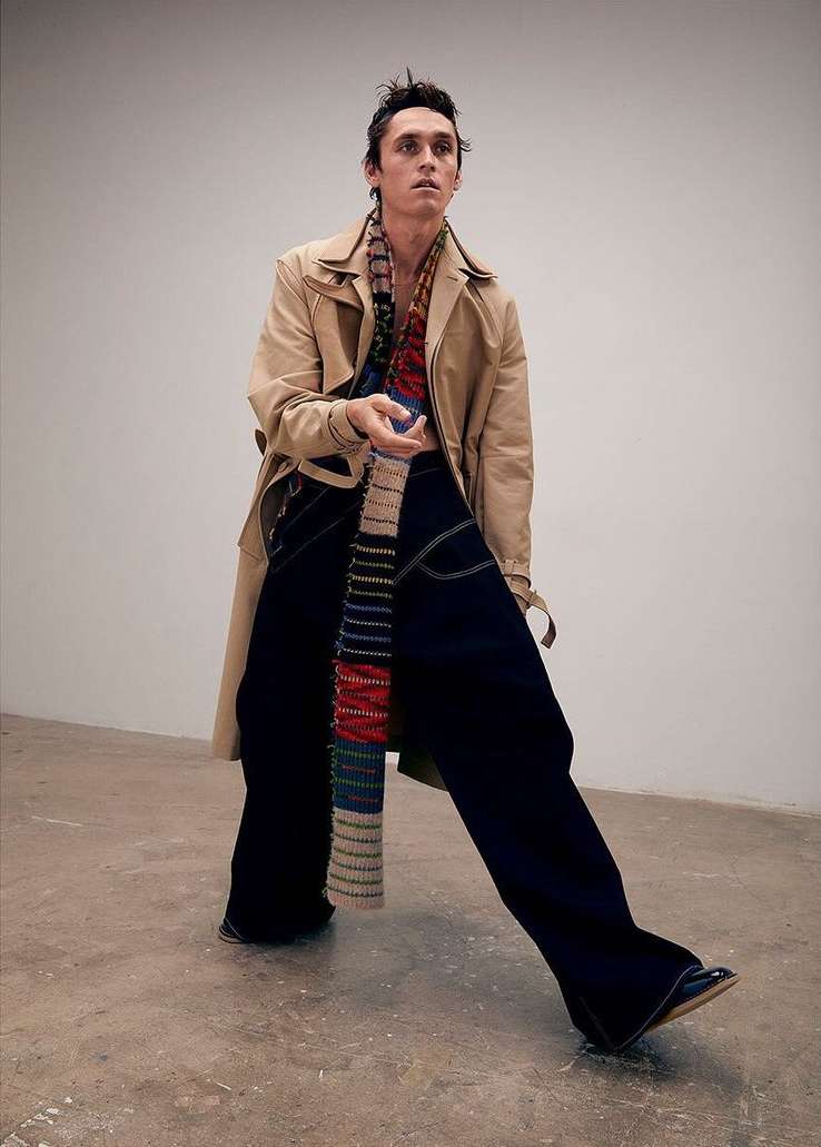 Fashionable adult male posing in a tan trench coat, dark pants, and striped scarf in a neutral indoor setting with studio lighting.