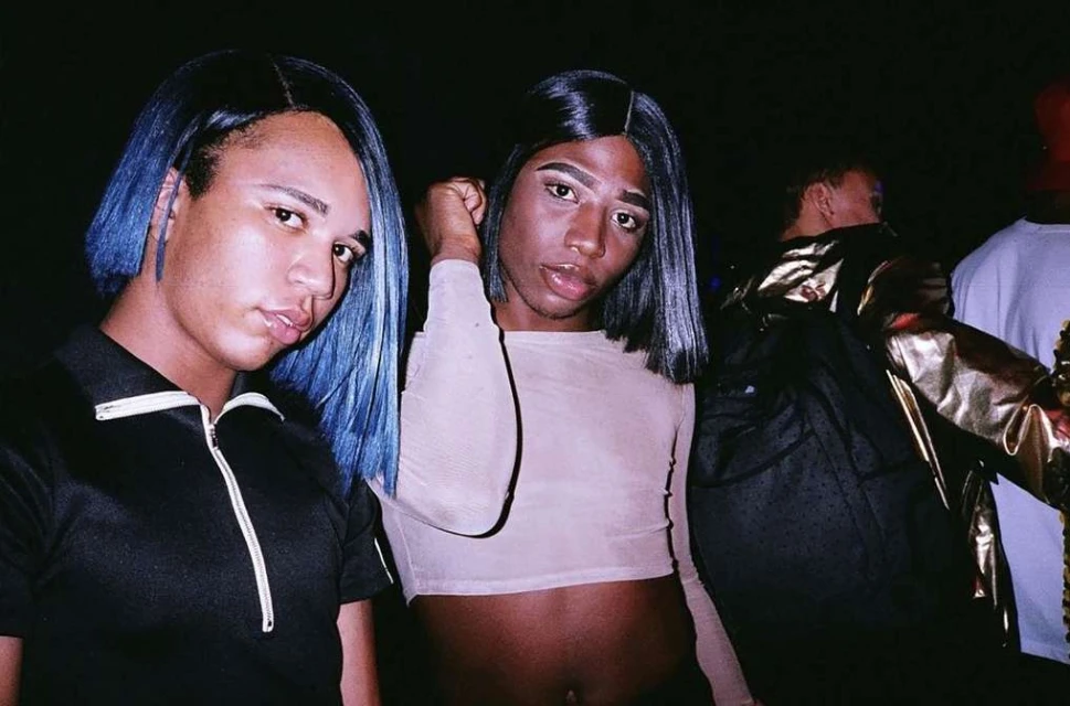 Two people posing together in an indoor setting with dark lighting, one with blue hair and a zip-up shirt, the other in a crop top.