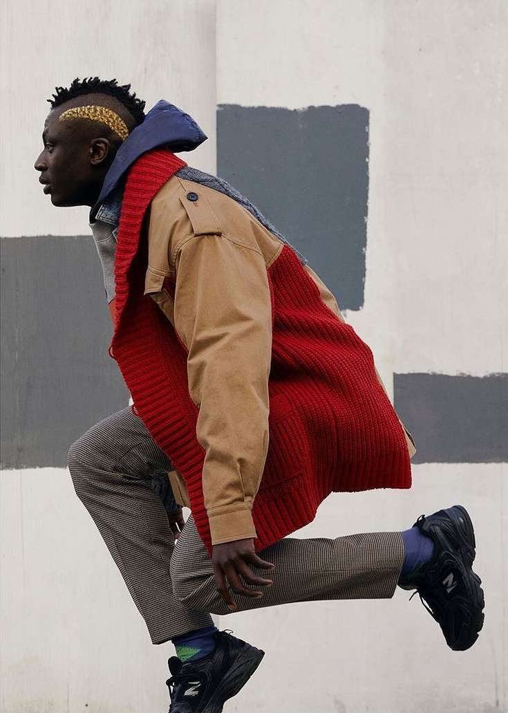A male model in motion wearing a multi-textured coat, striped pants, and black New Balance sneakers poses against a block painted wall.