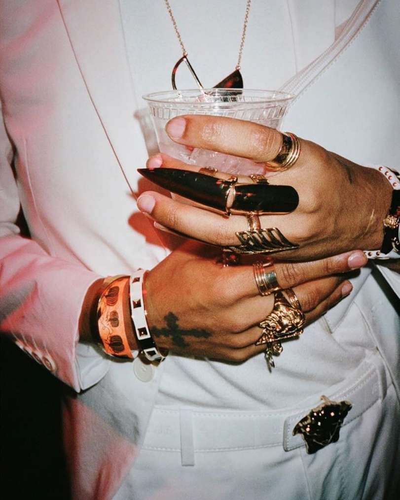 Close-up of hands adorned with multiple rings, bracelets, and a necklace, holding a clear plastic cup, wearing a white suit.
