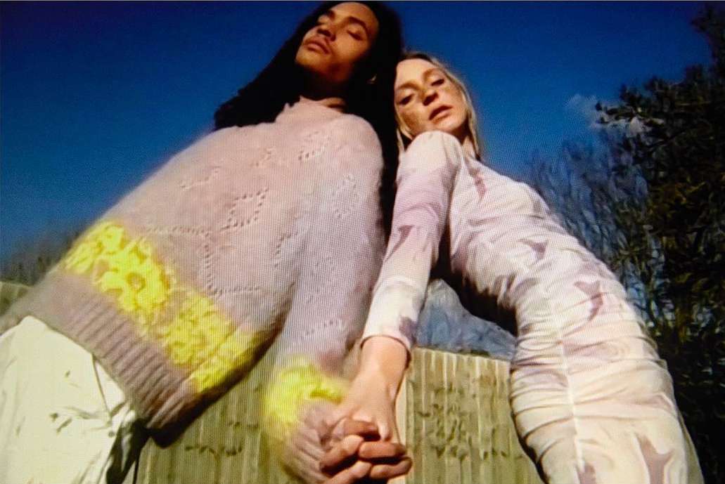 A stylish couple poses outdoors under a blue sky, featuring a man in a knit sweater and a woman in a patterned dress.