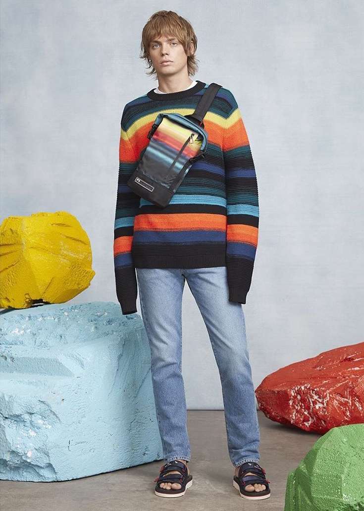 Male fashion model wears a striped sweater, crossbody bag, jeans, and sandals in a studio setting with colorful prop rocks.