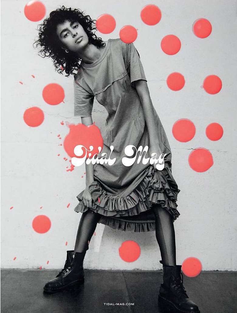 Fashion editorial shot of a woman with curly hair in a gray dress and black boots against a wall with coral dots.