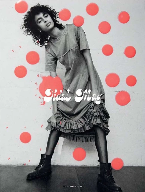 Fashion editorial shot of a woman with curly hair in a gray dress and black boots against a wall with coral dots.