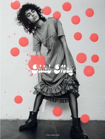 Fashion editorial shot of a woman with curly hair in a gray dress and black boots against a wall with coral dots.