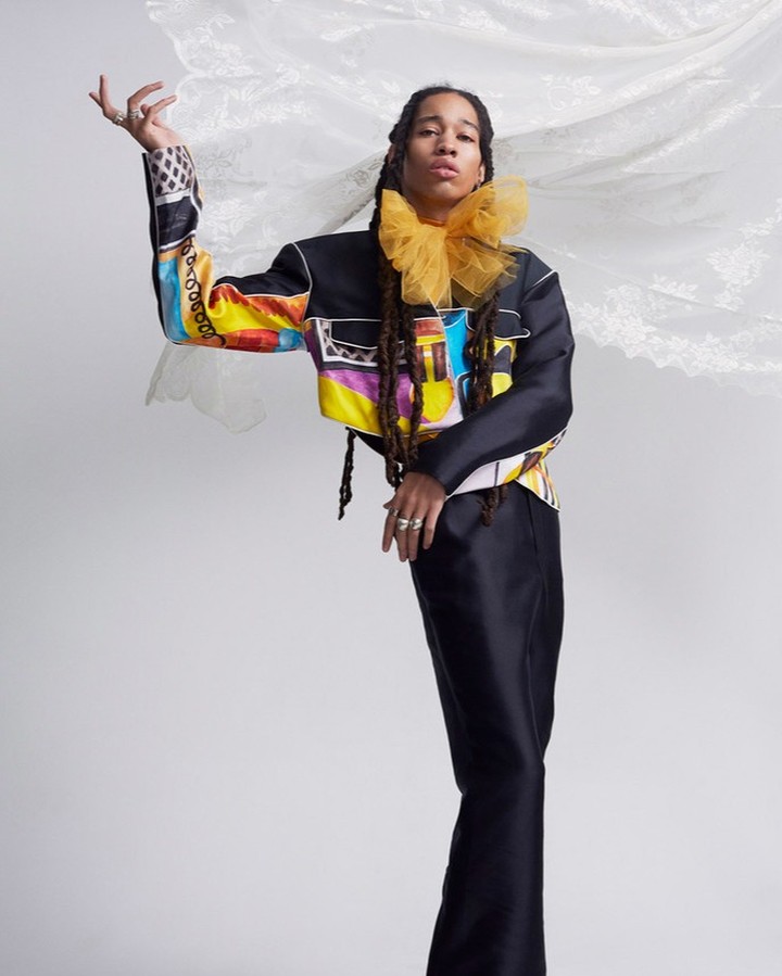 Model in a colorful patterned jacket and black pants poses in a studio with flowing sheer white fabric behind her, showcasing high fashion.