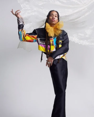 Model in a colorful patterned jacket and black pants poses in a studio with flowing sheer white fabric behind her, showcasing high fashion.