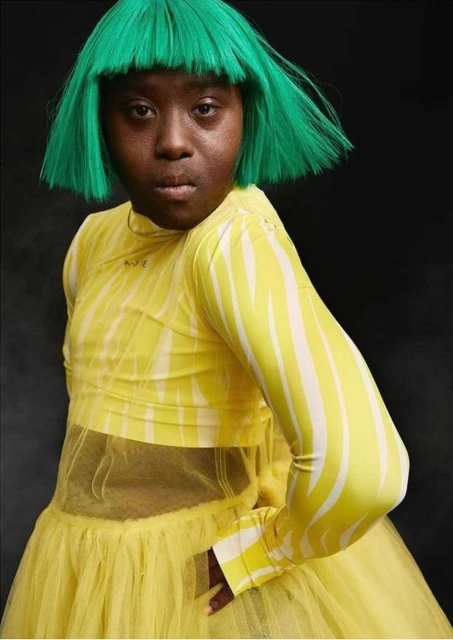 Woman with green hair and yellow dress posing against a dark background in a fashion portrait with studio lighting and a direct gaze.