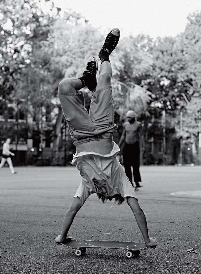 A person does a handstand on a skateboard in a black and white outdoor shot with other people and trees in the background.