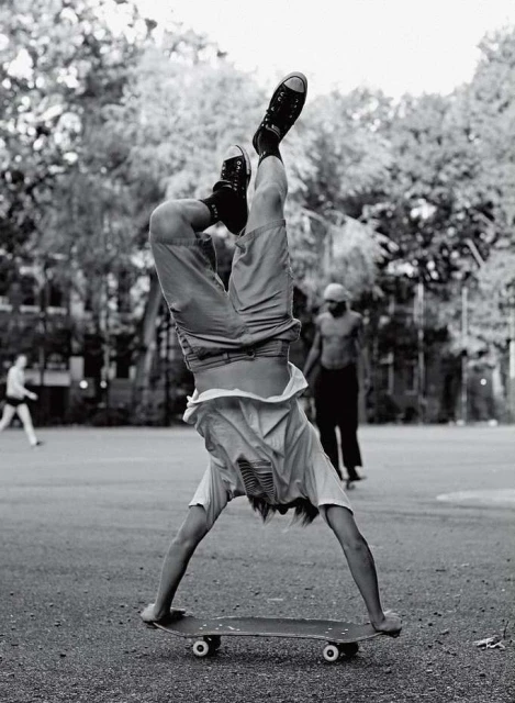A person does a handstand on a skateboard in a black and white outdoor shot with other people and trees in the background.