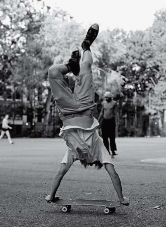 A person does a handstand on a skateboard in a black and white outdoor shot with other people and trees in the background.