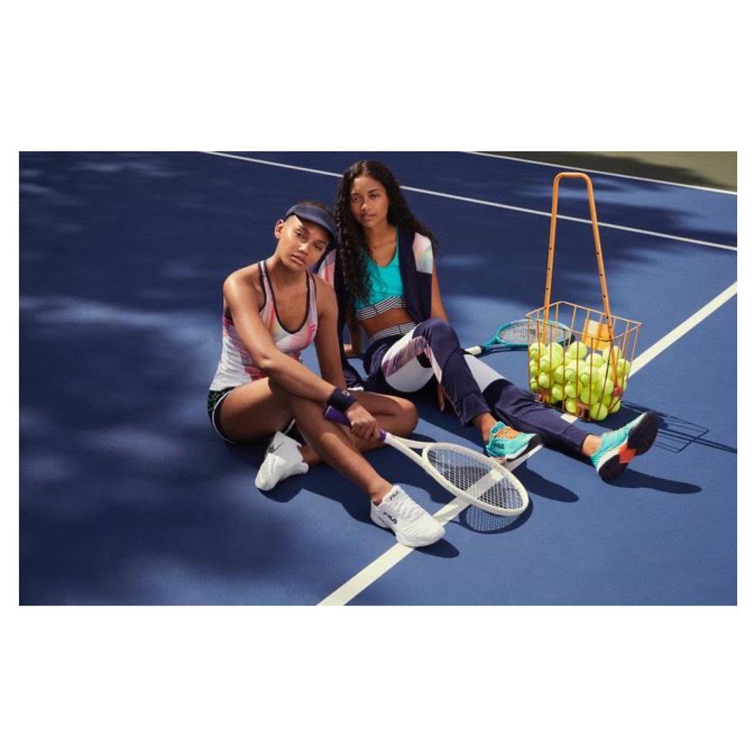 Two women sit on a blue tennis court with a basket of tennis balls. They are wearing athletic clothing and holding rackets.