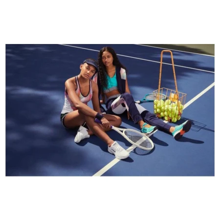 Two women sit on a blue tennis court with a basket of tennis balls. They are wearing athletic clothing and holding rackets.