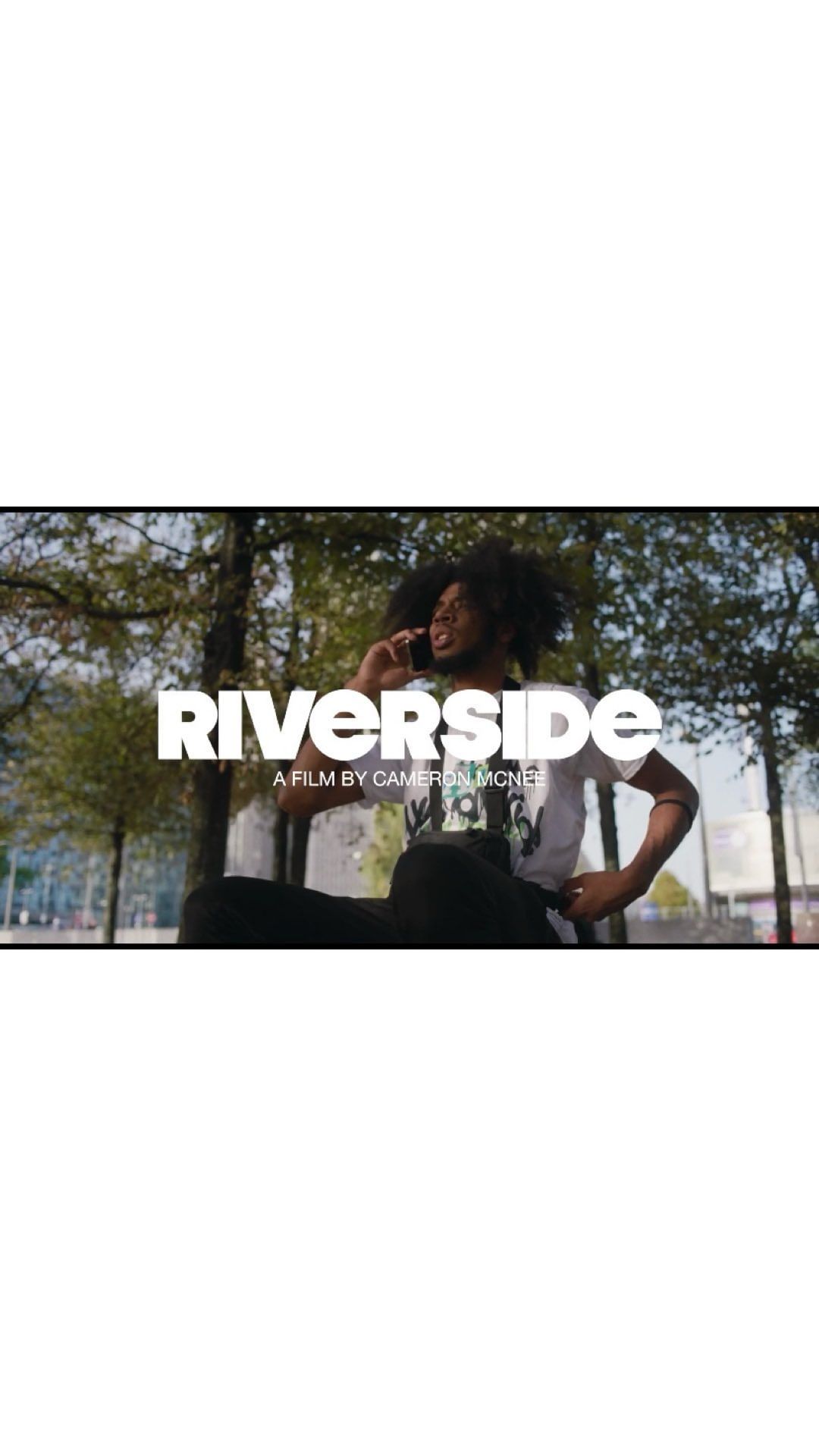 An adult male with dark hair is using a cellphone outside in an urban environment with green trees as the background. The word 'Riverside' is superimposed.