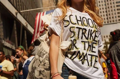 Woman wearing a white t-shirt with black handwritten text and carrying a canvas tote bag on a crowded city street.